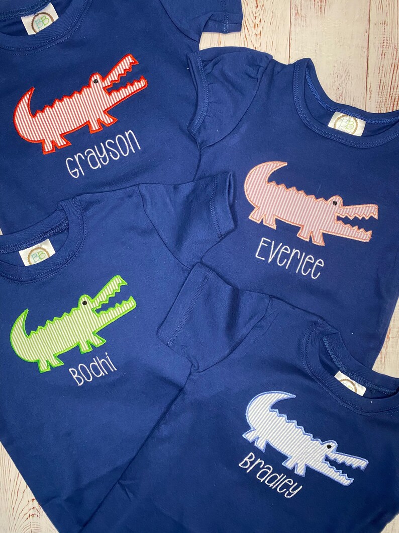 May include: Four navy blue t-shirts with alligator designs and personalised names. The alligators are red and white, pink and white, green and white, and blue and white. The names are Grayson, Everlee, Bodhi, and Bradley.