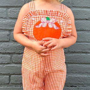 May include: A young child wearing a white and orange gingham jumpsuit with a large orange pumpkin appliqu&eacute; on the front. The pumpkin has a pink bow and green stem.