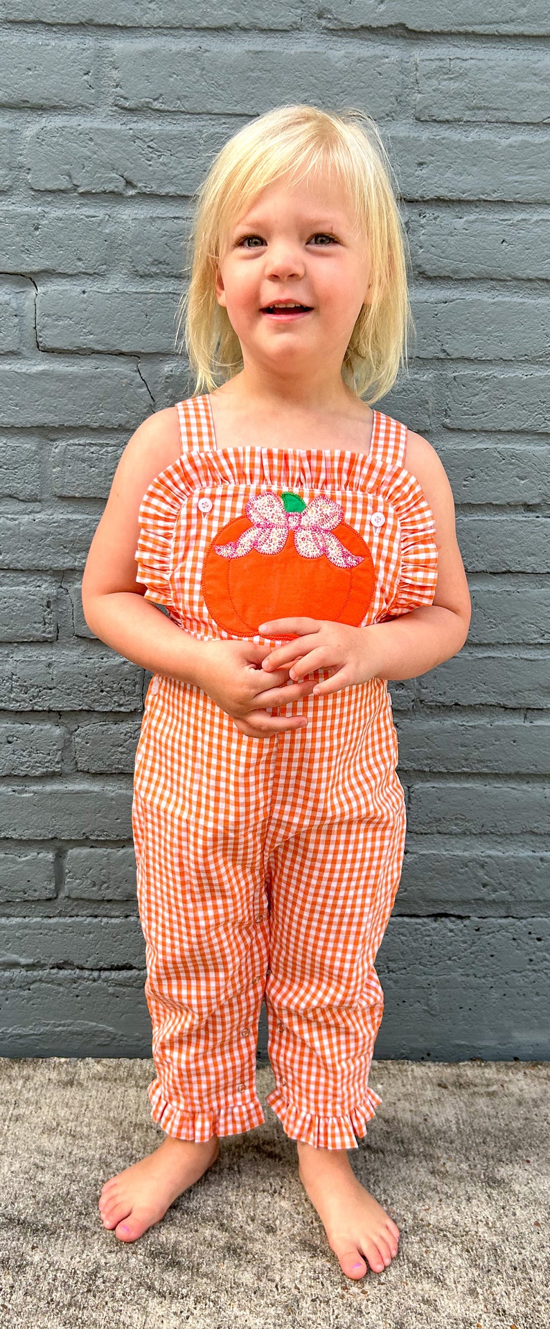 May include: A young child wearing a white and orange gingham jumpsuit with a large orange pumpkin appliqu&eacute; on the front. The pumpkin has a pink bow and green stem.