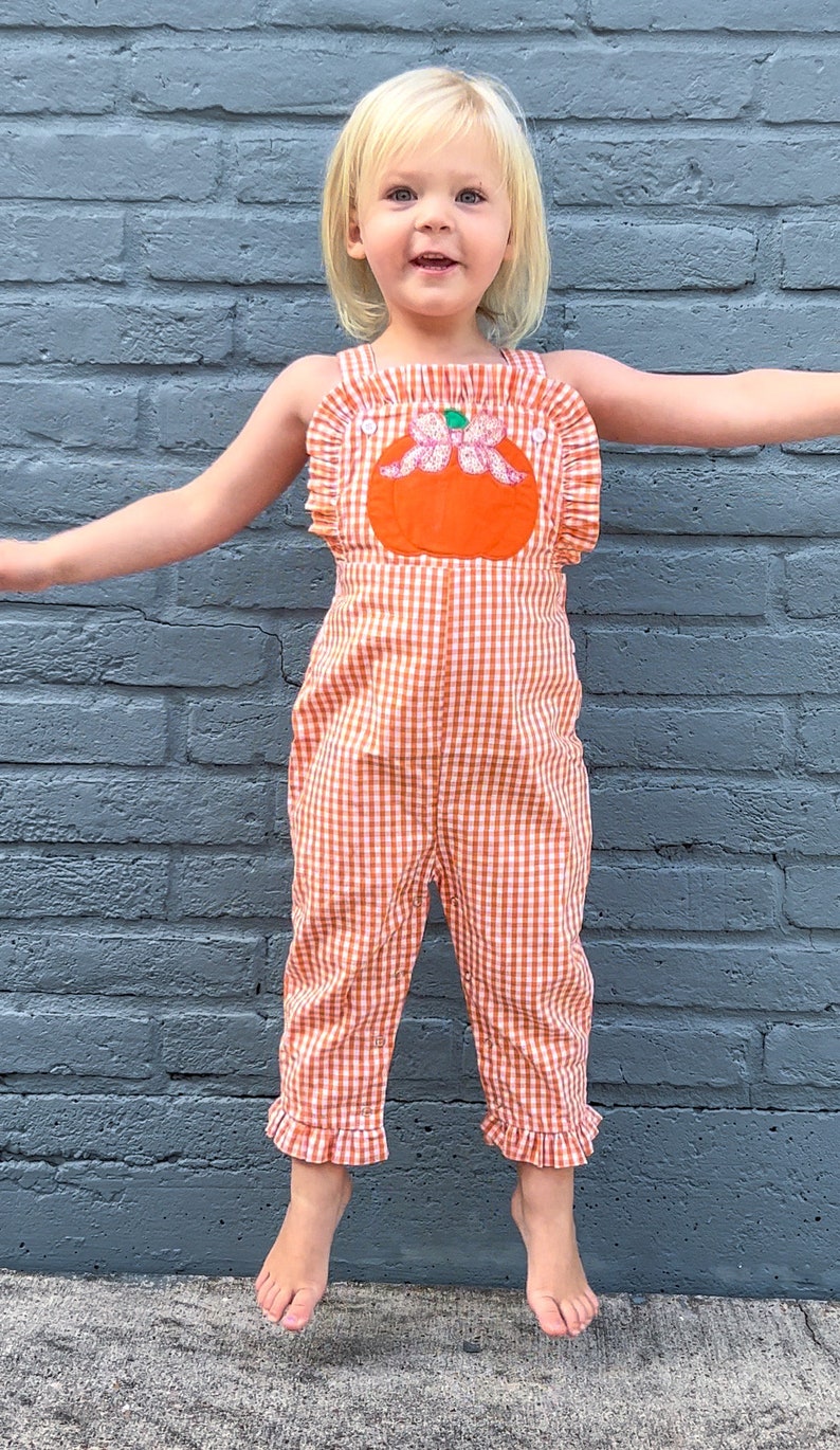 May include: A young girl wearing a sleeveless orange and white gingham jumpsuit with a pumpkin applique. The jumpsuit has ruffles around the neckline and the bottom of the legs.