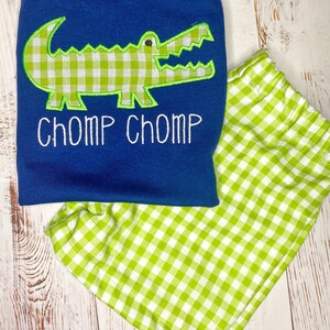 May include: A blue and green gingham baby outfit with a green alligator applique and the text "CHOMP CHOMP" embroidered on the shirt.