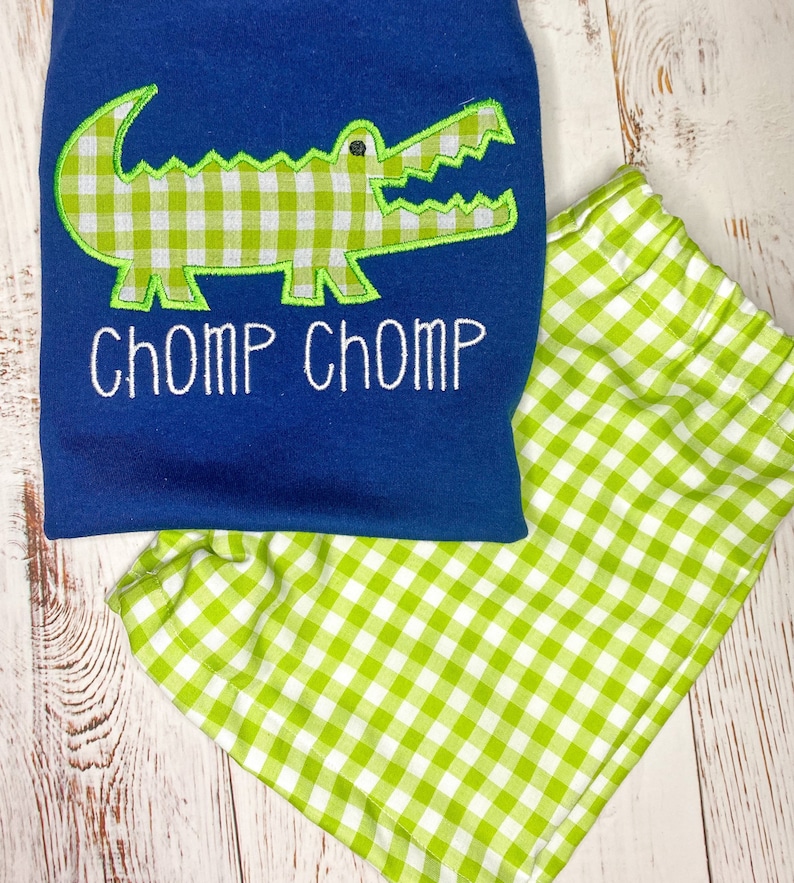 May include: A blue and green gingham baby outfit with a green alligator applique and the text "CHOMP CHOMP" embroidered on the shirt.