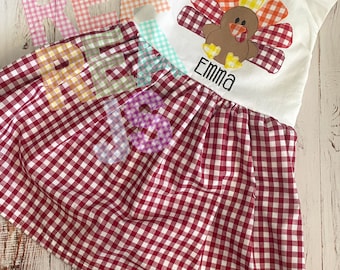 May include: A white sleeveless dress with a red and white gingham skirt. A brown turkey with a red, orange, and yellow gingham tail is appliqued on the bodice. The name "Emma" is embroidered below the turkey.