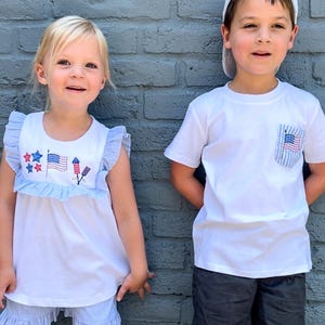 May include: Two children wearing patriotic themed clothing. A girl is wearing a white sleeveless shirt with blue and red stars, a red, white, and blue flag, and fireworks embroidered on it. She is also wearing blue and white striped shorts. A boy is wearing a white t-shirt with a red, white, and blue flag in the pocket. He is wearing black shorts and a white baseball cap.