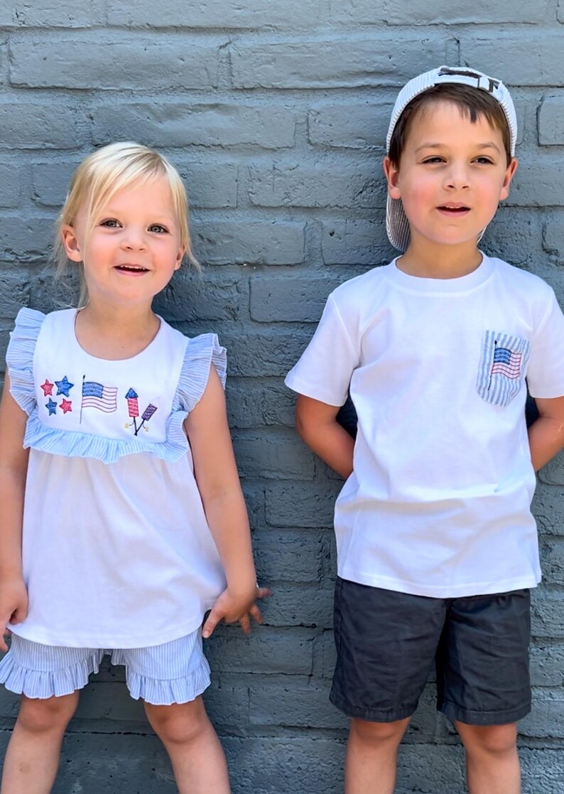 May include: Two children wearing patriotic themed clothing. A girl is wearing a white sleeveless shirt with blue and red stars, a red, white, and blue flag, and fireworks embroidered on it. She is also wearing blue and white striped shorts. A boy is wearing a white t-shirt with a red, white, and blue flag in the pocket. He is wearing black shorts and a white baseball cap.