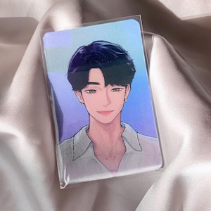 May include: A clear plastic card featuring a digital illustration of a person with dark hair and a white shirt. The illustration has a soft, colorful background with blue and purple hues. The card is displayed on a satin surface.