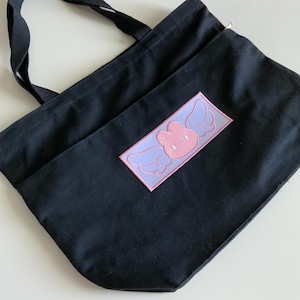 K&ouml;nnte beinhalten: Eine schwarze Canvas-Tragetasche mit einem rosa gestickten Aufn&auml;her eines Hasen mit Fl&uuml;geln. Der Aufn&auml;her hat eine rechteckige Form mit einem wei&szlig;en Umriss.
