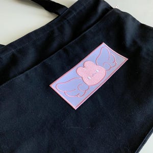 K&ouml;nnte beinhalten: Eine schwarze Tragetasche mit einem rosa und violetten gestickten Aufn&auml;her eines Kaninchens mit Fl&uuml;geln. Der Aufn&auml;her hat die Form eines Rechtecks.