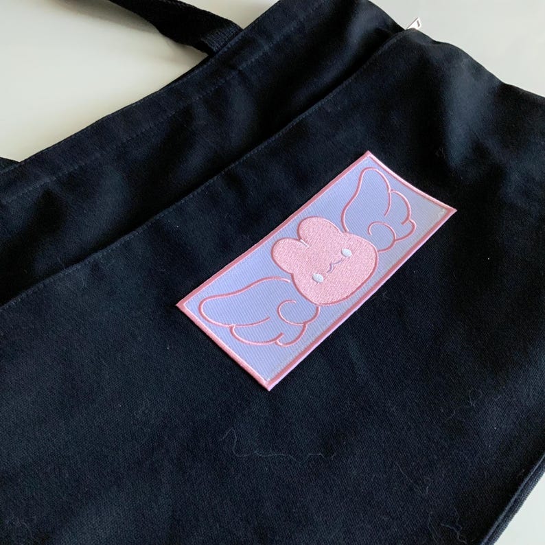K&ouml;nnte beinhalten: Eine schwarze Tragetasche mit einem rosa und violetten gestickten Aufn&auml;her eines Kaninchens mit Fl&uuml;geln. Der Aufn&auml;her hat die Form eines Rechtecks.