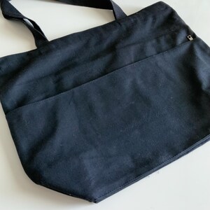 K&ouml;nnte beinhalten: Eine schwarze Canvas-Tragetasche mit einer Rei&szlig;verschlusstasche auf der Vorderseite. Die Tasche hat zwei Henkel.
