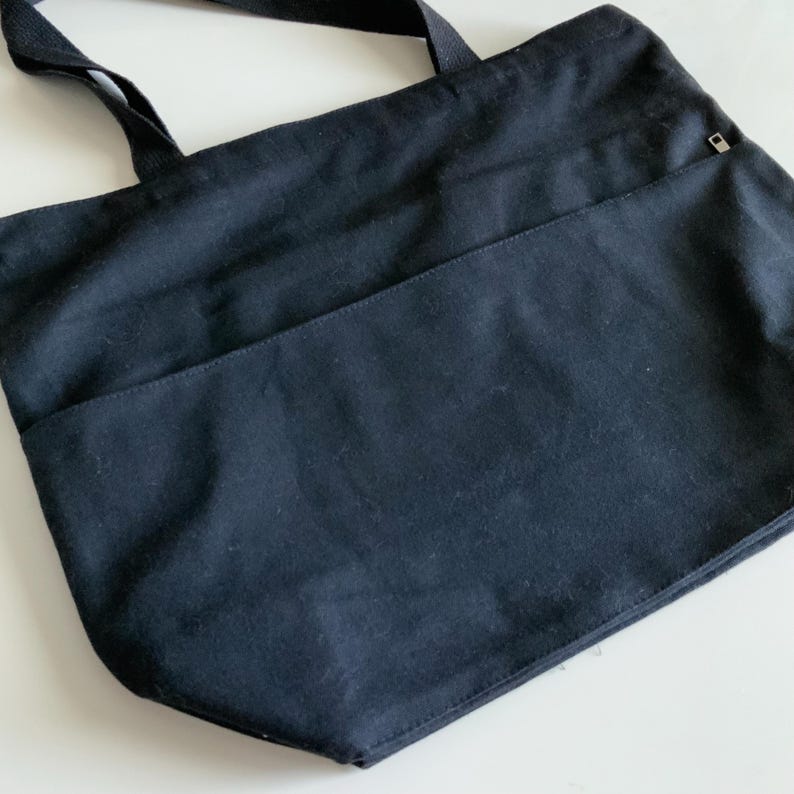 K&ouml;nnte beinhalten: Eine schwarze Canvas-Tragetasche mit einer Rei&szlig;verschlusstasche auf der Vorderseite. Die Tasche hat zwei Henkel.