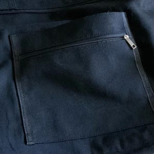 K&ouml;nnte beinhalten: Eine schwarze Canvas-Tasche mit einer Rei&szlig;verschlusstasche auf der R&uuml;ckseite. Der Rei&szlig;verschluss ist silberfarben und hat einen Schieber.