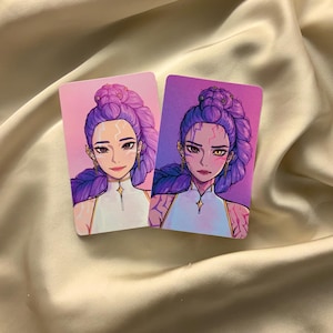 May include: Two rectangular cards featuring illustrations of a woman with purple hair styled in a bun. The woman wears a white top with a star-shaped pendant. The background gradients from pink to purple.