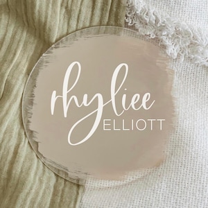 May include: A round, clear acrylic sign with a light brown background and white brushstrokes. The name "Rhylee Elliott" is written in white cursive script.