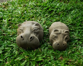 Puede incluir: Dos cabezas de hipopótamo de cerámica gris sobre un lecho de hierba verde.
