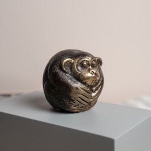 May include: A small, bronze-colored monkey figurine with a detailed, textured surface. The monkey is curled up in a seated position with its head resting on its arms.