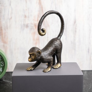 May include: A bronze monkey ornament with a curled tail and outstretched arm. The monkey has a dark brown body with gold accents on its feet and face. The ornament is on a grey block, with a green clock in the background.