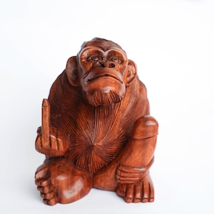 Peut inclure: Une sculpture en bois d'un singe assis, la main levée en signe d'insulte. Le singe est sculpté dans une couleur brun rougeâtre et a un visage détaillé.
