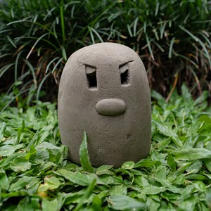 Może przedstawiać: Szara, zaokrąglona rzeźba ceramiczna z twarzą. Twarz ma dwa oczy, nos i usta. Rzeźba stoi na ł&oacute;żku z zielonej trawy.