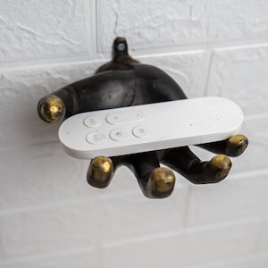 Könnte beinhalten: Eine weiße Fernbedienung mit den Tasten "OFF", "ON", "+", "-", "M" und "N" wird in einem dekorativen handförmigen Halter gehalten.