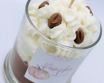 May include: A clear glass candle with a brown base and white whipped cream-like wax, topped with brown coffee bean-shaped wax pieces. A label reads "Pumpkin Spice Latte".