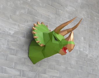 May include: A green and gold papercraft dinosaur head mounted on a gray brick wall. The dinosaur has three horns and an open mouth.