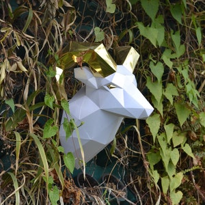 Pode incluir: Escultura de papel branco de uma cabeça de carneiro com detalhes dourados. A escultura está montada em uma parede verde coberta de videiras.