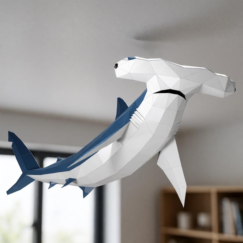 May include: A low-poly papercraft hammerhead shark sculpture featuring a geometric white and blue design. The shark is depicted in a swimming pose with a wide head, a dark blue dorsal fin, and a matching blue tail fin, constructed from flat panels.