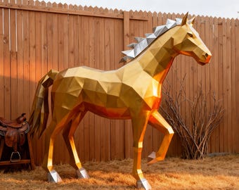 May include: A large, geometric golden horse sculpture with silver accents on the mane and hooves. The sculpture is outdoors, set against a wooden fence and dry grass, with a saddle visible in the background.