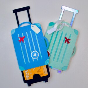 May include: Two decorative suitcase-shaped gift card holders. One is blue with a red airplane and white stripes, the other is mint green with a red airplane and white stripes. Each has a pull-out drawer.