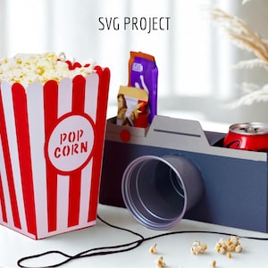 May include: A red and white striped popcorn container with the word "POP CORN" printed on it, a camera-shaped cardboard box with a can inside, and a black cord.