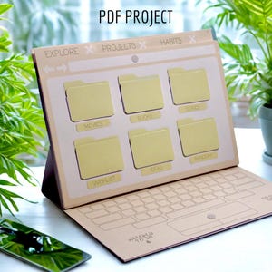 May include: A beige paper organizer designed to resemble a laptop screen, featuring labeled file folders for movies, books, series, ideas, random, and a wishlist. The text "PDF PROJECT" is at the top. A smartphone rests nearby.