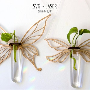 Puede incluir: Dos maceteros de pared de madera con forma de mariposa, cada uno con un vial de vidrio con una planta verde. Las alas de madera están cortadas con láser con detalles intrincados. El texto "SVG - LASER 3mm & 1/8" es visible.