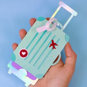 May include: A light teal paper suitcase with a red airplane and heart-shaped sticker. The suitcase has a handle and a ribbon. The suitcase is held in a hand against a light blue background.