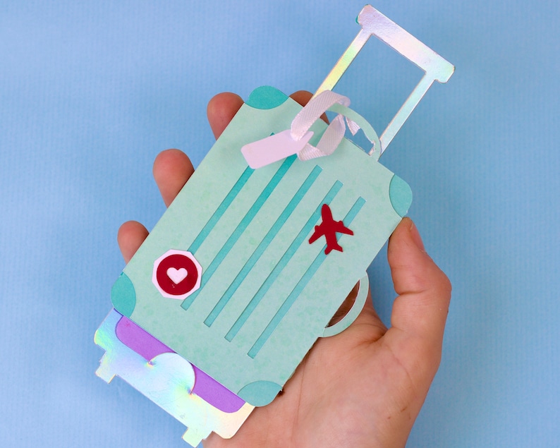 May include: A light teal paper suitcase with a red airplane and heart-shaped sticker. The suitcase has a handle and a ribbon. The suitcase is held in a hand against a light blue background.
