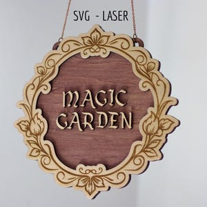 May include: A circular wooden hanging sign featuring the text MAGIC GARDEN in raised lettering. The sign has a dark brown center and a light tan decorative border with floral engravings. The text SVG - LASER is visible at the top.