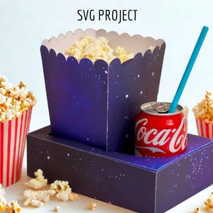 Puede incluir: Un conjunto de palomitas de maíz y refrescos con un tema cósmico. El recipiente de palomitas es azul marino con puntos blancos en forma de estrella y un borde festoneado. Una lata de Coca-Cola roja con una pajita azul se encuentra sobre una base a juego.