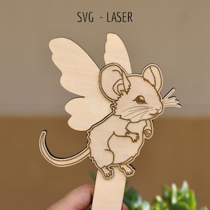 Puede incluir: Un recorte de madera de color claro de un ratón con alas de hada, con detalles grabados del rostro, bigotes y cuerpo. El texto SVG - LASER está impreso en la parte superior. El recorte está unido a un palo de madera vertical en la base.