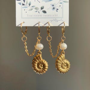 May include: Gold-toned earrings with a seashell design. Each earring features a small pearl and a chain connecting the top hoop to the seashell pendant. The earrings are displayed on a white card with text that reads "Your review means a lot."