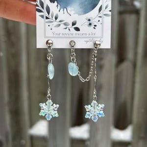 Op de afbeelding: Zilverkleurige oorbellen met sneeuwvlokhangers en lichtblauwe edelsteenaccenten. De oorbellen zijn verbonden door een fijne ketting. De achtergrond toont een kaart met de tekst "Your review means a lot".