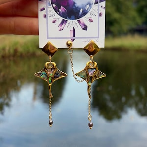 May include: Gold-toned earrings featuring a stylized ray design with iridescent abalone shell inlays. Each earring has a diamond-shaped top and a dangling chain with a dark gray pearl. The earrings are connected by a delicate chain.