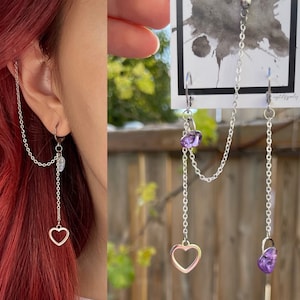 May include: Silver chain earrings featuring a heart-shaped charm and a dangling crystal. One earring connects to the ear with a chain that goes up to the ear. The other earring has a purple crystal and a chain.