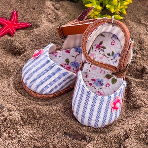 Puede incluir: Un par de zapatos de bebé a rayas azules y blancas con detalles florales y correas de cuero marrón. Los zapatos están sobre una superficie arenosa.