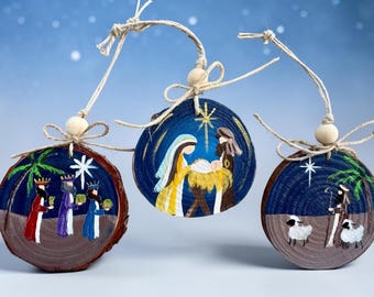 May include: Three circular wood slice ornaments featuring hand-painted nativity scenes. Each piece hangs from a natural twine loop with a wooden bead. The designs depict the three wise men, the holy family, and a shepherd with sheep under a star.