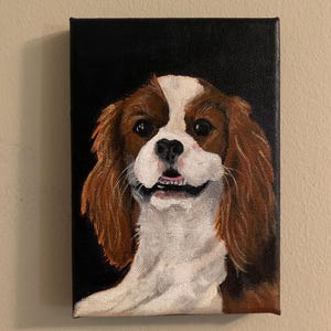 May include: A rectangular canvas painting of a Cavalier King Charles Spaniel dog. The dog has brown and white fur, with a black background. The dog is smiling, with its mouth slightly open, and the painting is mounted on a wall.