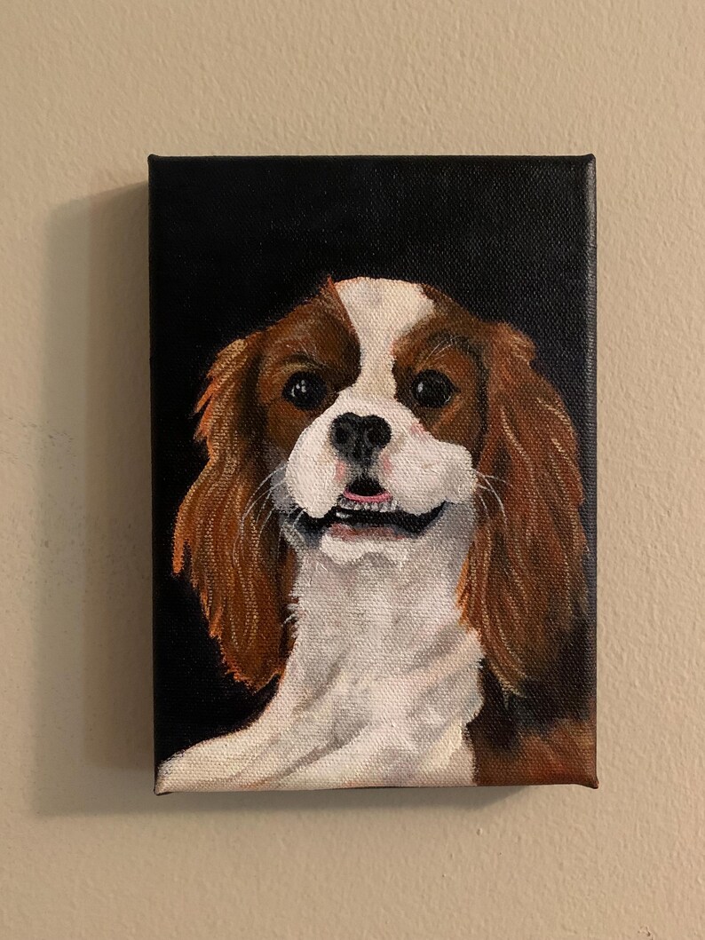 May include: A rectangular canvas painting of a Cavalier King Charles Spaniel dog. The dog has brown and white fur, with a black background. The dog is smiling, with its mouth slightly open, and the painting is mounted on a wall.
