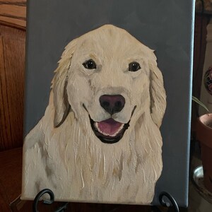 May include: A canvas painting of a Golden Retriever dog with a happy expression. The dog has cream-colored fur, a pink nose, and a pink tongue. The background is a solid gray color. The painting is displayed on a black metal easel.