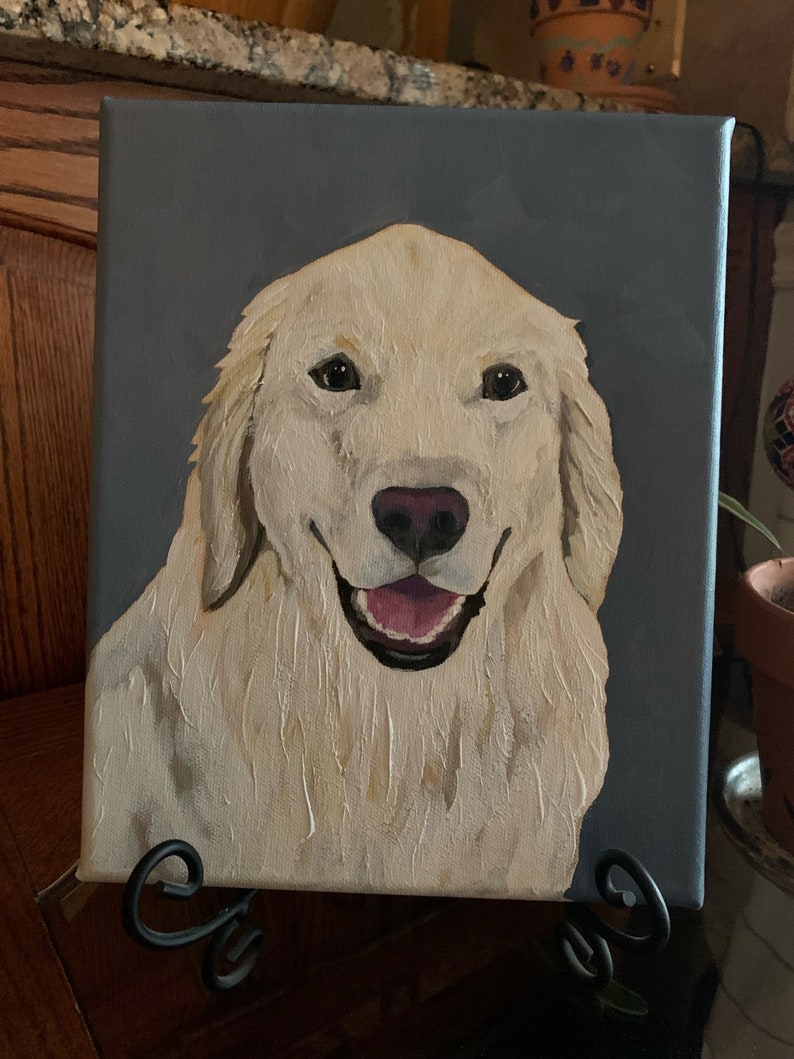 May include: A canvas painting of a Golden Retriever dog with a happy expression. The dog has cream-colored fur, a pink nose, and a pink tongue. The background is a solid gray color. The painting is displayed on a black metal easel.