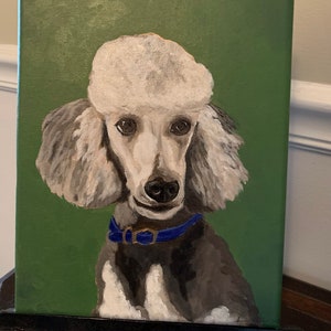 May include: A painted portrait of a poodle on a green background. The poodle has white fur with gray accents, a blue collar, and a black nose. The painting is displayed on a black easel.