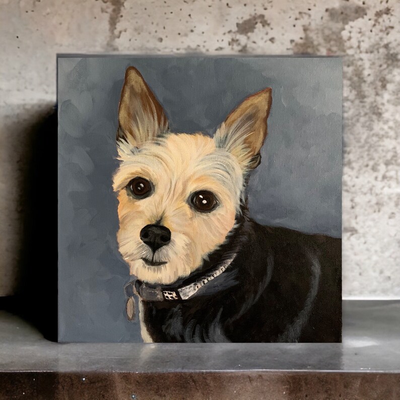May include: Square canvas painting of a small dog with a cream and black coat, brown eyes, and large ears. The dog wears a collar with a tag. The background is a solid, muted blue-gray color. The painting is displayed on a gray surface.